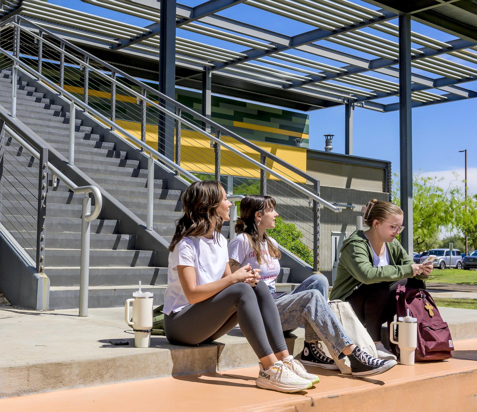 Three students are sitting on the steps outside a campus building, appearing relaxed and engaged in conversation in a casual outdoor setting.