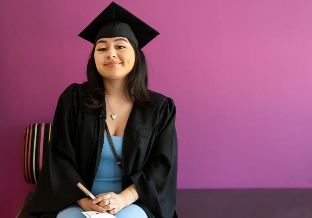 Female student graduate wearing graduation cap and gown, posing in front of purple painted wall.