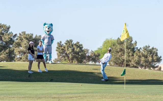 Two women and Codi, the mascot, watch a man play golf